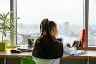 Woman writing at desk with a view of the city skyline through a large window.